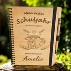 Personalisiertes Einschulungsbuch aus Holz mit Wunschnamen Amelie, Regenbogen- und Schultüten-Gravur, schwarzer Spiralbindung und Holz-Kugelschreiber.