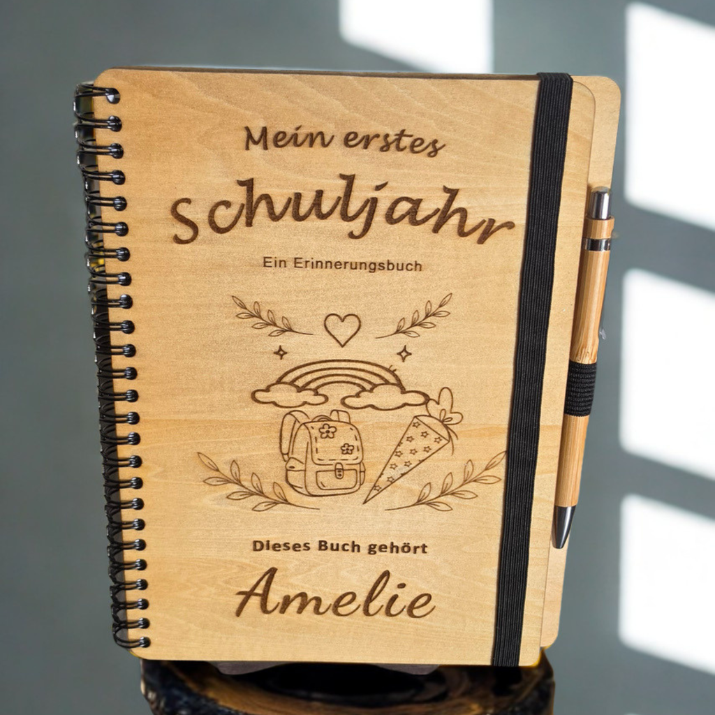 Personalisiertes Einschulungsbuch aus Holz mit Wunschnamen Amelie, Regenbogen- und Schultüten-Gravur, schwarzer Spiralbindung und Holz-Kugelschreiber.