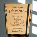 Personalisiertes Einschulungsbuch aus Holz mit Wunschnamen Amelie, Regenbogen- und Schultüten-Gravur, schwarzer Spiralbindung und Holz-Kugelschreiber.