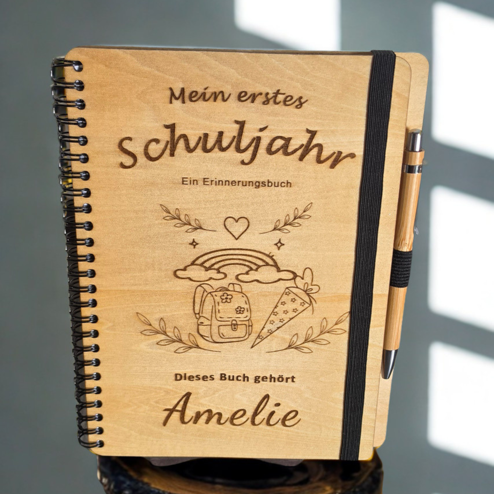 Personalisiertes Einschulungsbuch aus Holz mit Wunschnamen Amelie, Regenbogen- und Schultüten-Gravur, schwarzer Spiralbindung und Holz-Kugelschreiber.