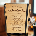 Personalisiertes Einschulungsbuch aus Holz mit Wunschnamen Amelie, Regenbogen- und Schultüten-Gravur, schwarzer Spiralbindung und Holz-Kugelschreiber.