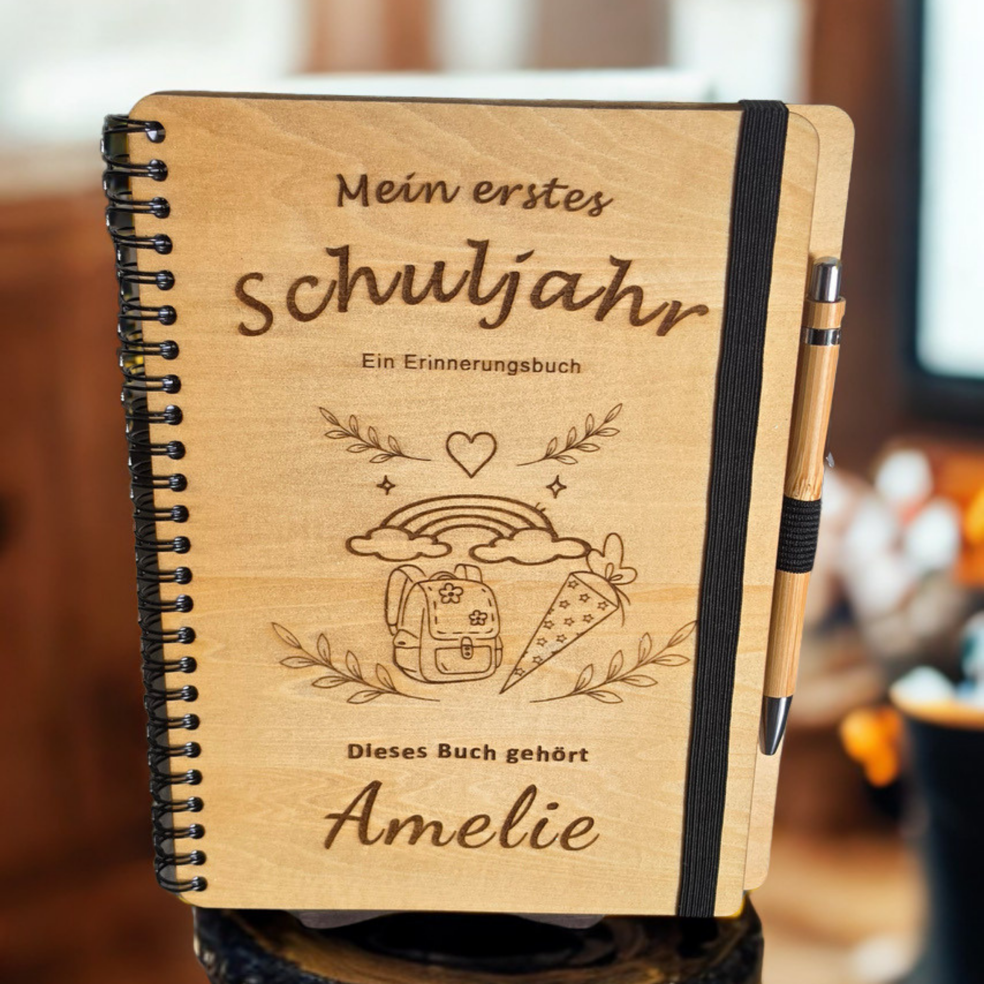 Personalisiertes Einschulungsbuch aus Holz mit Wunschnamen Amelie, Regenbogen- und Schultüten-Gravur, schwarzer Spiralbindung und Holz-Kugelschreiber.