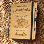 Personalisiertes Einschulungsbuch aus Holz mit Wunschnamen Amelie, Regenbogen- und Schultüten-Gravur, schwarzer Spiralbindung und Holz-Kugelschreiber.