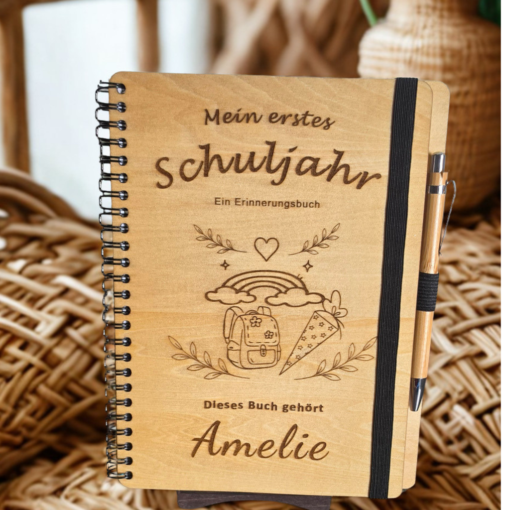 Personalisiertes Einschulungsbuch aus Holz mit Wunschnamen Amelie, Regenbogen- und Schultüten-Gravur, schwarzer Spiralbindung und Holz-Kugelschreiber.