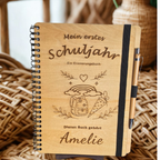 Personalisiertes Einschulungsbuch aus Holz mit Wunschnamen Amelie, Regenbogen- und Schultüten-Gravur, schwarzer Spiralbindung und Holz-Kugelschreiber.