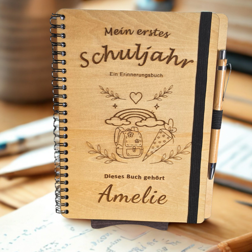 Personalisiertes Einschulungsbuch aus Holz mit Wunschnamen Amelie, Regenbogen- und Schultüten-Gravur, schwarzer Spiralbindung und Holz-Kugelschreiber.