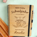 Personalisiertes Einschulungsbuch aus Holz mit Wunschnamen Amelie, Regenbogen- und Schultüten-Gravur, schwarzer Spiralbindung und Holz-Kugelschreiber.
