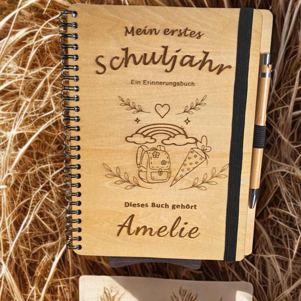 Personalisiertes Einschulungsbuch aus Holz mit Wunschnamen Amelie, Regenbogen- und Schultüten-Gravur, schwarzer Spiralbindung und Holz-Kugelschreiber.