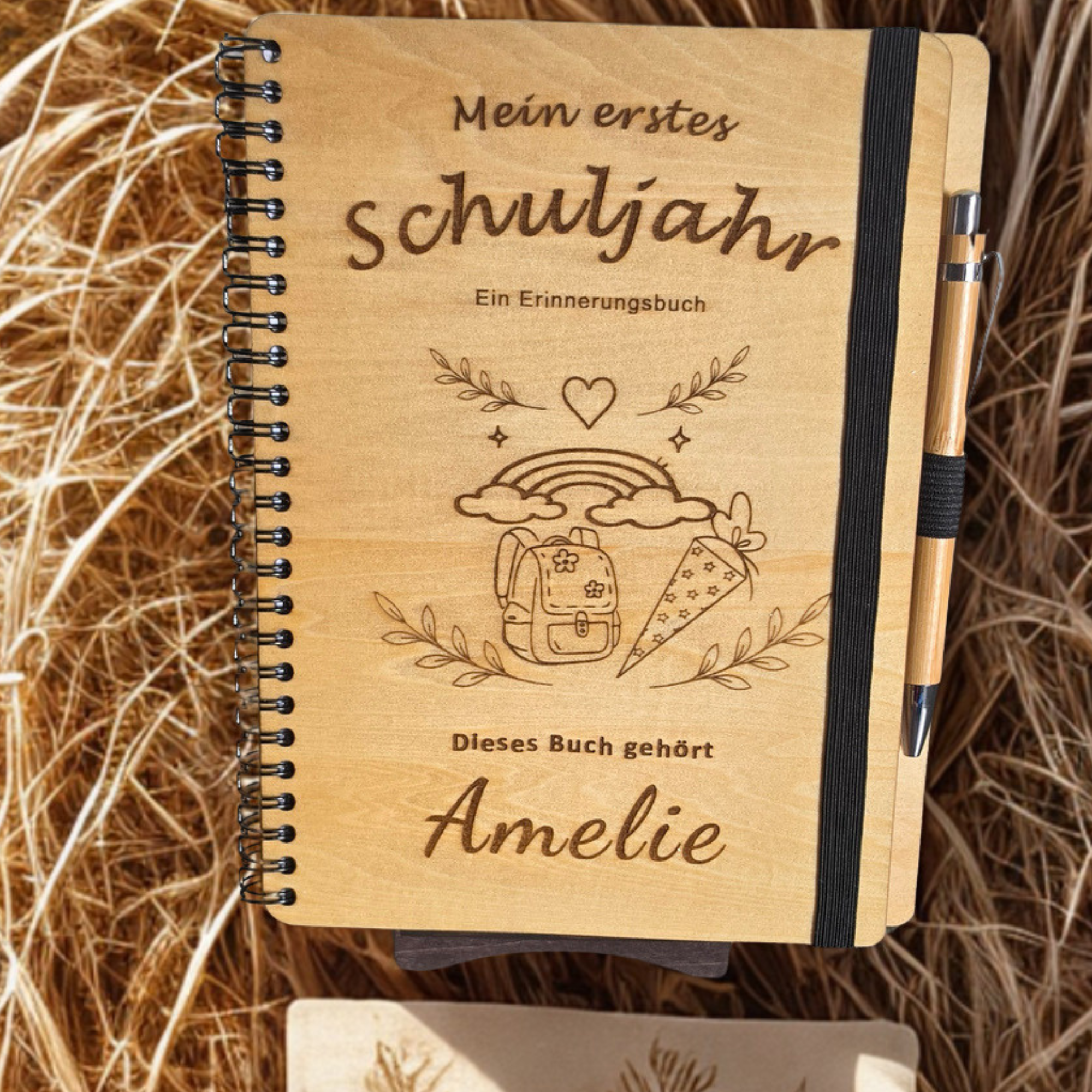 Personalisiertes Einschulungsbuch aus Holz mit Wunschnamen Amelie, Regenbogen- und Schultüten-Gravur, schwarzer Spiralbindung und Holz-Kugelschreiber.